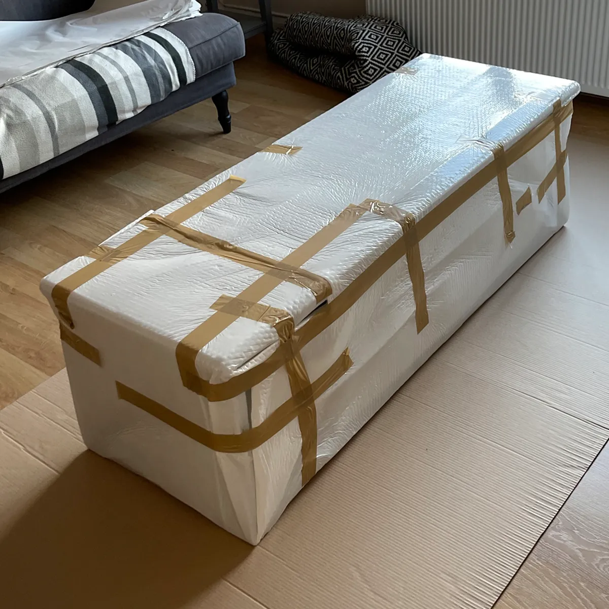 Large item wrapped in protective white packing material and secured with tape, sitting on cardboard floor protection in a living room before a move