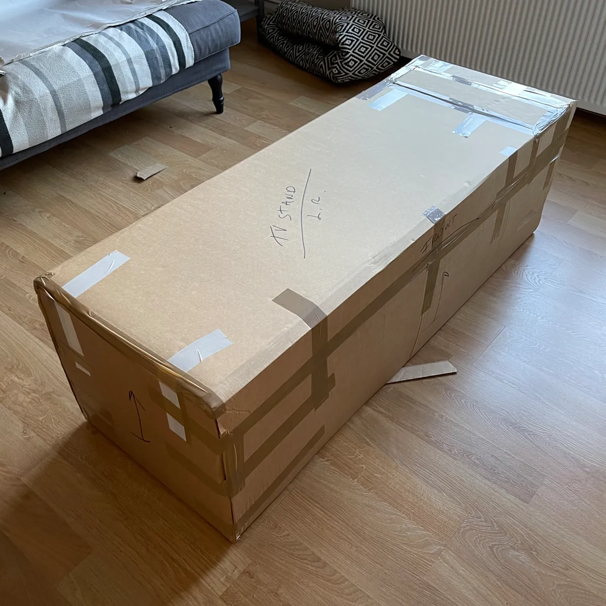 Large cardboard shipping box taped shut and labeled by hand, resting on a wooden floor in a living room next to a sofa and radiator